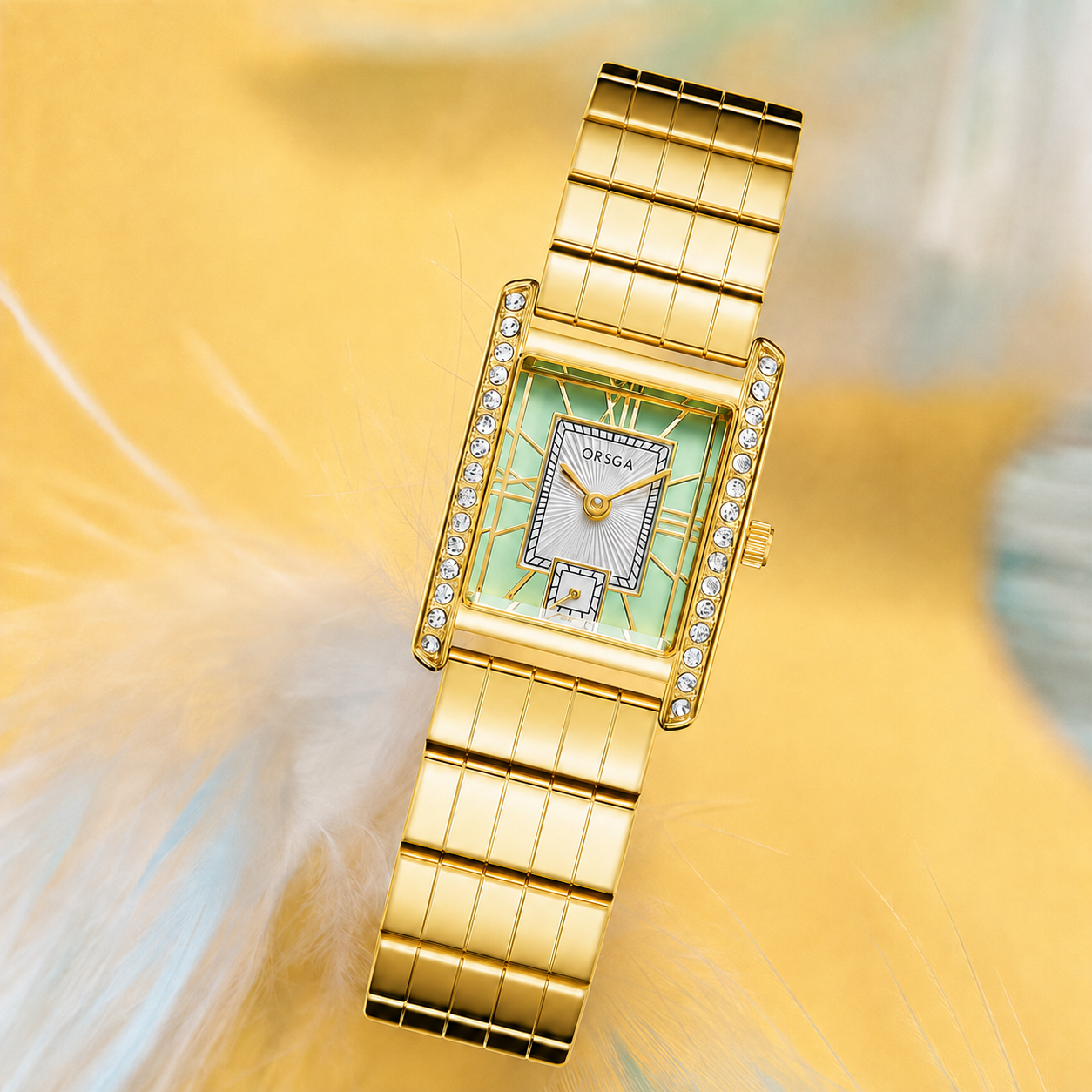 A gold-tone rectangular watch with a light green dial, crystal rhinestone accents around the bezel, and a matching gold-tone link bracelet, shown on a soft yellow background with blurred feathers.
