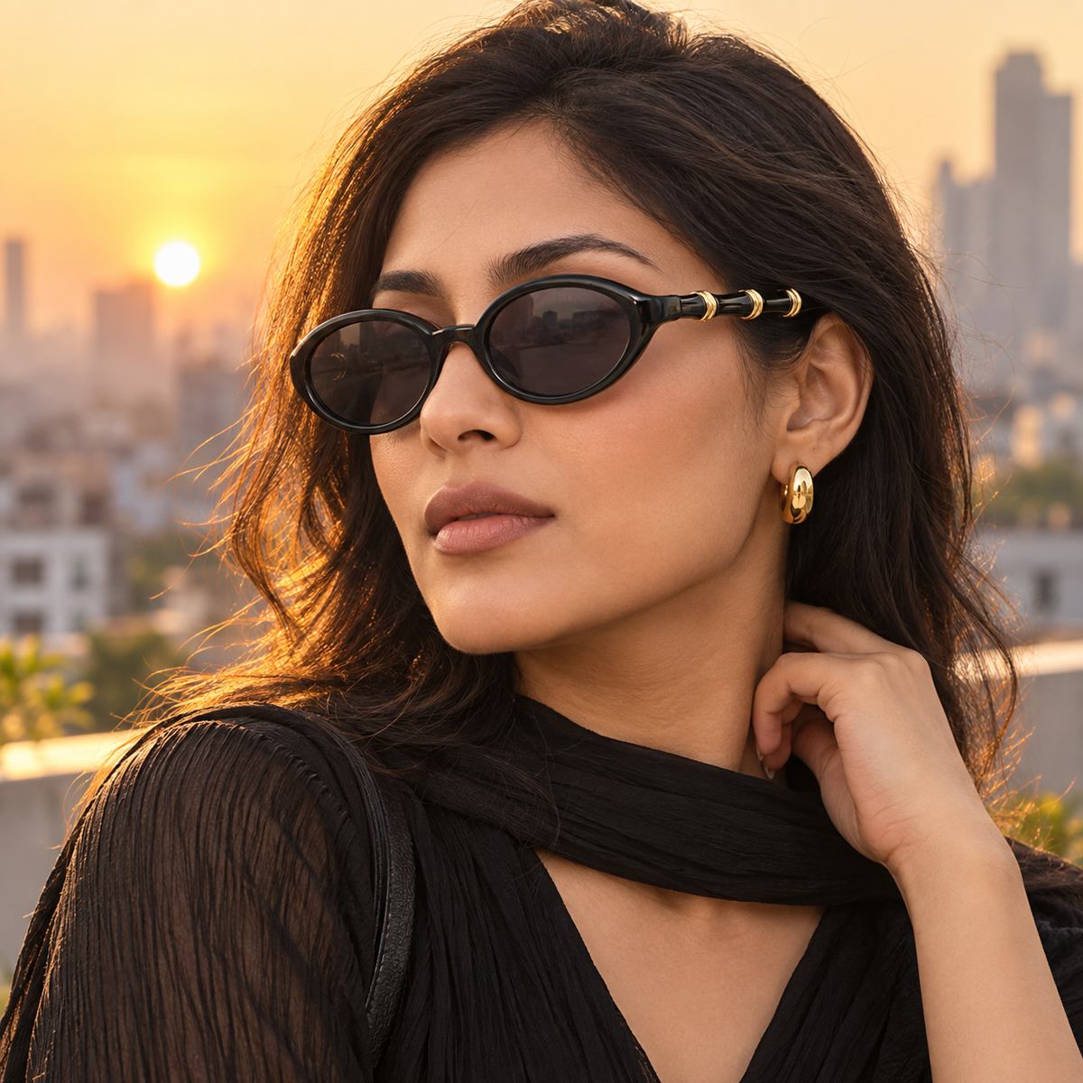A woman wearing black oval sunglasses with gold metal accents on the temples, photographed outdoors against a warm sunset city skyline.