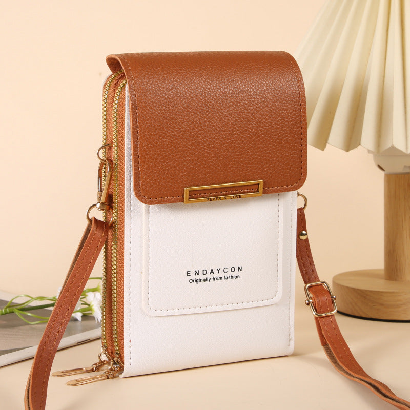 A small crossbody phone bag with a brown pebbled faux-leather top flap, a white front pocket, and gold-tone hardware, shown standing upright on a beige tabletop with a lamp in the background.
