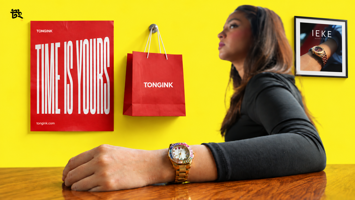 A woman sits at a wooden table against a bright yellow wall, wearing a gold-tone Tongink watch. Red Tongink shopping bags and a red poster reading 'TIME IS YOURS' are hanging on the wall, along with a framed IEKE watch advertisement.